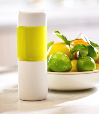 A white bottle with yellow liquid inside stands on a counter next to a bowl of fresh lemons and limes.