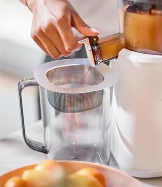 A hand operates a juicer, pouring juice through a metal strainer into a clear pitcher on a kitchen counter.