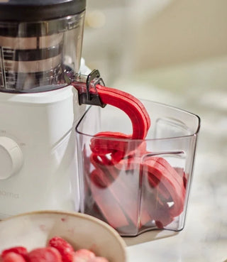 A white juicer extrudes bright red fruit pulp into a clear plastic container; a bowl of cut strawberries is visible in the foreground.