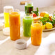 Three On-the-Go Glass Bottles filled with green and orange juice rest on a marble counter, accompanied by a white bottle and a plate of assorted fruits in the background. Designed for convenient juice transport, each bottle has its white cap placed nearby. Bright sunlight illuminates the scene, emphasizing the freshness and practicality of these sleek On-the-Go Glass Bottles—an ideal choice for your healthy lifestyle.
