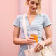 A woman in a light blue dress holds juice in the On-the-Go Glass Bottle, featuring a silicone sleeve, and places it into a beige tote bag while standing in front of a pink background.