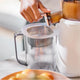 A hand operates a juicer, pouring juice through the Pulp Strainer into a clear pitcher for easy, pulp-free juicing. A bowl of fruit sits on the table in the foreground.