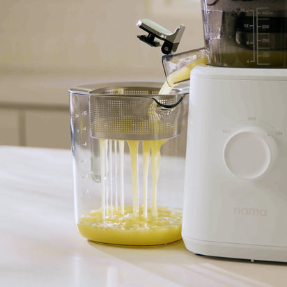 A white Nama juicer pours yellow juice through the Pulp Strainer into a clear container on a kitchen counter. The juicer features a visible dial and uses the Pulp Strainer for efficient, no-pulp juicing.