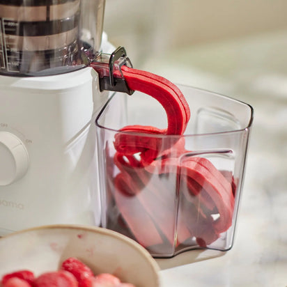 A juicer ejects red fruit pulp into a clear container, with a bowl of strawberries nearby on a light-colored surface—perfect ingredients for making homemade sorbet using the Sorbet Attachment and crafting a healthy dessert.