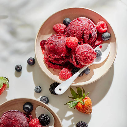 Two bowls of homemade sorbet, crafted using the Sorbet Attachment, are artfully served with generous scoops topped with fresh raspberries, blueberries, strawberries, and blackberries. Spoons are placed in each bowl, while a few berries are scattered on the white surface nearby for a deliciously healthy dessert.