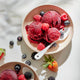 Two bowls of homemade sorbet, crafted using the Sorbet Attachment, are artfully served with generous scoops topped with fresh raspberries, blueberries, strawberries, and blackberries. Spoons are placed in each bowl, while a few berries are scattered on the white surface nearby for a deliciously healthy dessert.