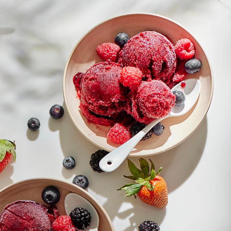 Two bowls of homemade sorbet, crafted using the Sorbet Attachment, are artfully served with generous scoops topped with fresh raspberries, blueberries, strawberries, and blackberries. Spoons are placed in each bowl, while a few berries are scattered on the white surface nearby for a deliciously healthy dessert.
