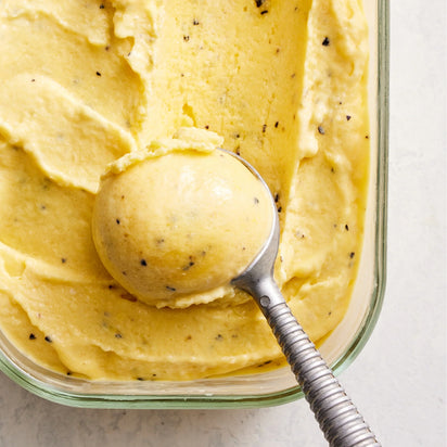 A metal ice cream scoop holds a rounded scoop of yellow ice cream with visible black specks, resting atop more homemade sorbet in a rectangular glass container—perfect for enjoying a healthy dessert made fresh with the Sorbet Attachment.