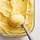 A metal ice cream scoop holds a rounded scoop of yellow ice cream with visible black specks, resting atop more homemade sorbet in a rectangular glass container—perfect for enjoying a healthy dessert made fresh with the Sorbet Attachment.