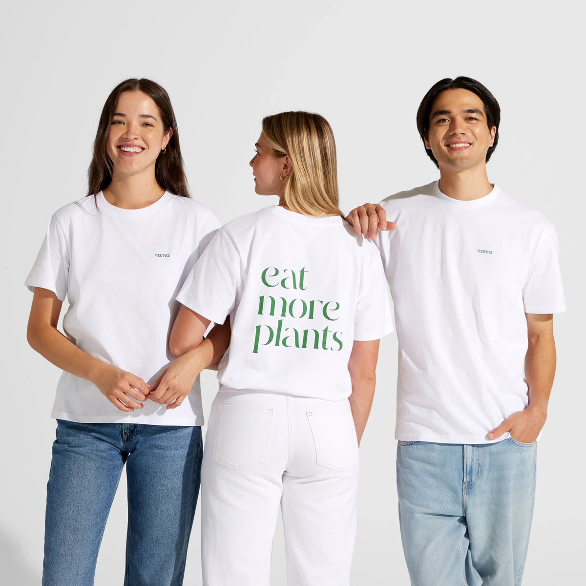 Three people in white t-shirts and jeans stand together. The person in the middle wears the Eat More Plants T-shirt, featuring bright green lettering on the back and made from 100% combed cotton for extra comfort. The other two are dressed in plain white relaxed fit tees. All are smiling.