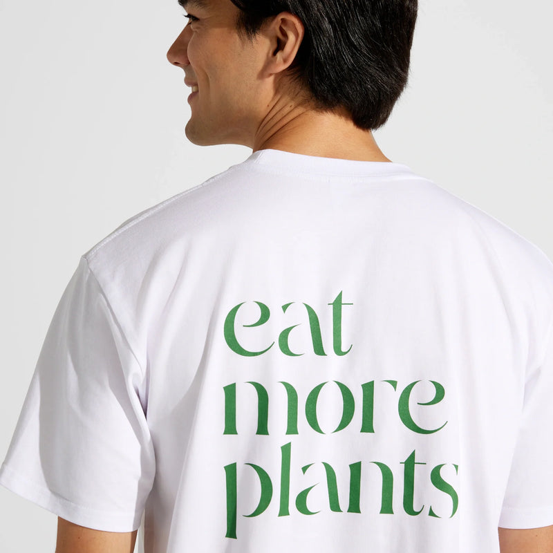 A smiling person faces slightly to the left, wearing the Eat More Plants T-shirt in white. The relaxed fit tee is made from 100% combed cotton and features green text on the back that reads “eat more plants.”.