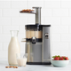 A silver and black Nama J1 Plug Type I cold press juicer sits on the counter with almonds being fed in. Nearby are a glass bottle and glass of almond milk, highlighting how easy it is to clean while making fresh, nutrient-rich juices.