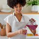 A woman in a light gray apron smiles as she pours vibrant purple juice made with the easy-to-clean Nama J1 Plug Type G cold press juicer into a glass in a modern kitchen.