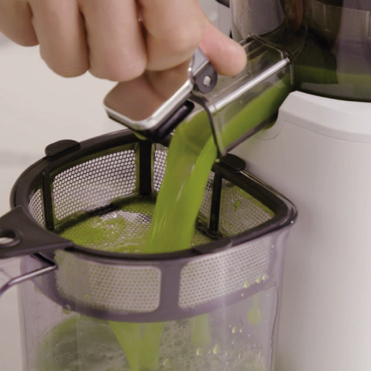 A hand pours bright green juice from a juicer spout into the Nama Pulp Strainer for J1 & C2 by Nama, which is set over a clear container.