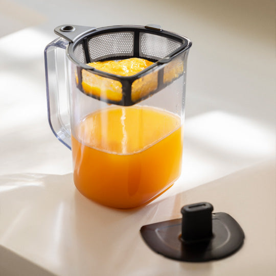 The Nama Pulp Strainer for J1 & C2 by Nama sits atop a clear plastic pitcher filled with orange juice and large orange halves. The lid rests nearby on a white surface, all lit by natural sunlight.