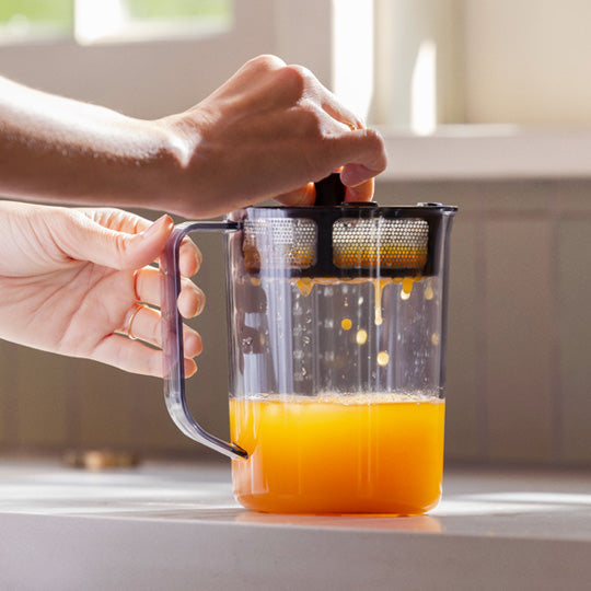 With sunlight streaming in, a person uses the Nama Pulp Strainer for J1 & C2 by Nama to strain fresh orange juice into a clear container on the kitchen counter.