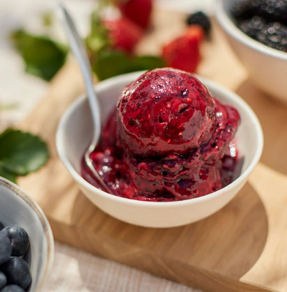 A bowl of dark red sorbet made with the Sorbet Attachment sits on a wooden board with a spoon. Fresh berries and an extra bowl of fruit sauce are blurred in the background.