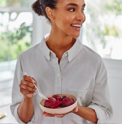 A woman in a light button-up shirt smiles as she enjoys homemade blueberry sorbet made with the Sorbet Attachment, eating with a spoon in a bright, airy room.