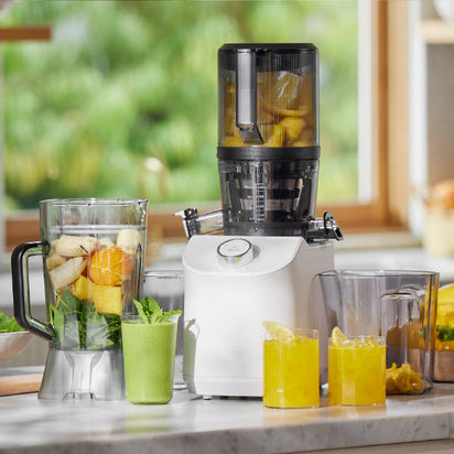 The Nama C2, a white multifunction juicer on a kitchen counter filled with chopped fruit, is surrounded by a blender of fruit, a green smoothie, two glasses of yellow juice with ice, and bowls of fresh fruit in a bright, sunlit kitchen.