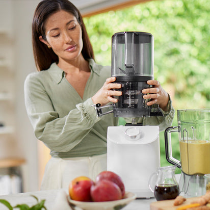 A woman in a light green blouse assembles the Nama C2 cold press juicer in a bright kitchen. Fresh fruits and a pitcher of green juice are on the counter, with large windows displaying lush greenery outside.