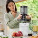 In a sunlit kitchen, a woman in a light green blouse assembles the white Nama C2 Plug Type G cold press juicer. On the countertop are a blender with green smoothie, apples, and a small jug. Greenery is visible outside the window.