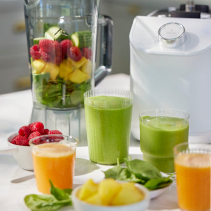 The Nama C2 juicer, filled with spinach, pineapples, cucumbers, and raspberries, sits beside its white base. In front are glasses of orange and green smoothies, with bowls of spinach and raspberries nearby.