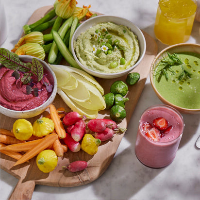 A wooden board with colorful vegetables, pink and green dips, and a bowl of green soup sits next to a glass of strawberry smoothie made with the Nama C2: Global Model (TESTING) blender on a marble surface.