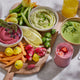 A wooden board of colorful veggies, dips, and zucchini flowers pairs perfectly with green soup made using the Nama C2 blender and a cold-pressed yellow drink plus a pink smoothie from the Nama C2 juicer.