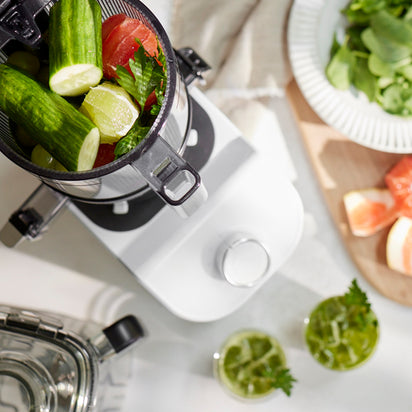 Top view of the Nama C2 cold press juicer filled with cucumbers, tomatoes, and leafy greens, surrounded by glasses of fresh juice, a cutting board with sliced fruit, and a plate of vegetables on a white countertop.