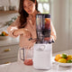 In a bright kitchen, a woman uses the Nama C2: Global Model (TESTING) juicer to extract fresh juice from whole fruits, pouring it into a pitcher while assorted citrus rests in a bowl on the counter.