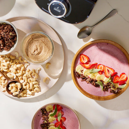 The Nama C2: Global Model (TESTING) blends a fresh smoothie bowl, topped with strawberries, banana, granola, and mint. Cashews, nut butter, a spoon, and a camera sit nearby in soft sunlight.