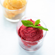 A glass cup with bright red sorbet—made from nutrient-rich juices using the Nama J1 Plug Type G cold press juicer—is topped with fresh mint. Another glass cup in the background contains yellow sorbet, slightly blurred.