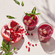 Two glasses of nutrient-rich pomegranate juice with ice and grape garnish sit beside a halved pomegranate, its seeds spilling out—perfect for enjoying fresh juice made in the Nama J1 cold press juicer.