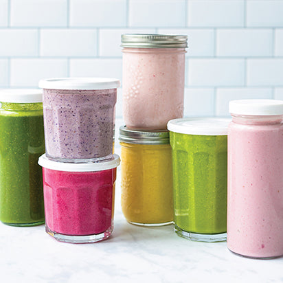 An array of vibrant smoothies in jars and bottles—green, pink, purple, yellow, magenta—displayed on a white countertop with tiled backsplash, highlights the science-based nutrition found in Blending for Beginners.