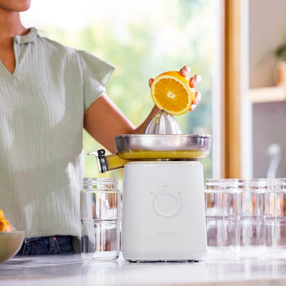 A person uses the Citrus Attachment to juice half an orange, with fresh juice pouring into a glass in a bright kitchen where clear glasses sit beside the juicer.