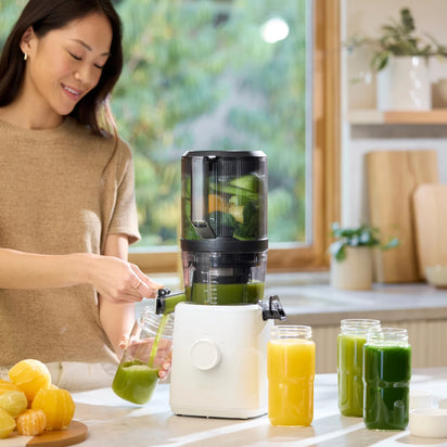 A woman smiles as she pours fresh green juice from a juicer into a glass jar in a bright kitchen. The Glass Bottle Set and halved citrus fruits sit on the counter, highlighting perfect storage solutions for homemade juice.