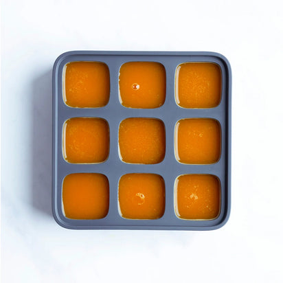 The Nama Ice Cube Tray, made of gray silicone with nine square compartments filled with orange liquid, is shown from above on a white surface.