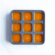 The Nama Ice Cube Tray, made of gray silicone with nine square compartments filled with orange liquid, is shown from above on a white surface.
