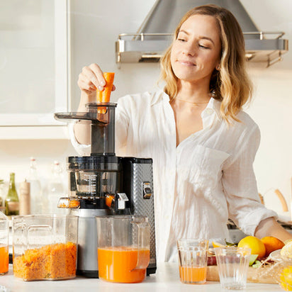 A woman in a white shirt smiles as she uses the Nama J1 cold press juicer, adding carrot pieces. Fresh juice and grated pulp are on the kitchen counter, surrounded by fruits and vegetables.