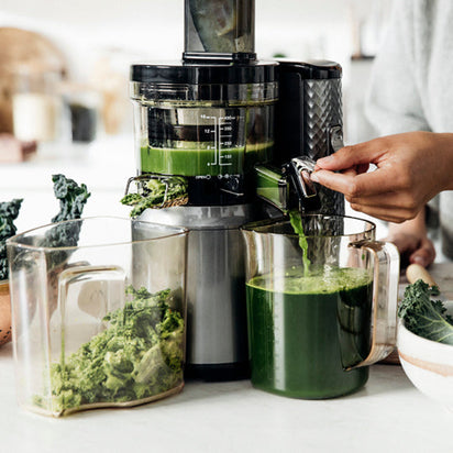 Using the Nama J1 Plug Type E juicer on a kitchen counter, someone extracts green juice from leafy vegetables, collecting juice in one container and pulp in another.