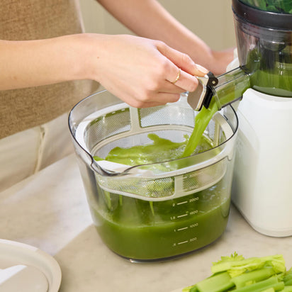 A person uses the Nama J2 XL to make green celery juice, which pours into a clear container with measurement markings. Celery stalks rest next to this easy-to-clean juicer on a white countertop.