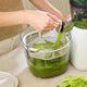 A person uses the Nama J2 XL to make green celery juice, which pours into a clear container with measurement markings. Celery stalks rest next to this easy-to-clean juicer on a white countertop.