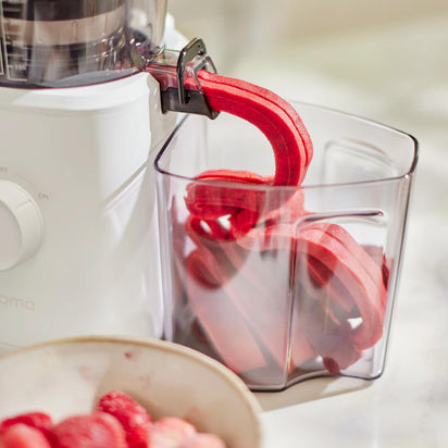 The Nama J2 XL cold press juicer expels vibrant red fruit pulp into a clear container, with a bowl of strawberries and raspberries beside it on a marble countertop.