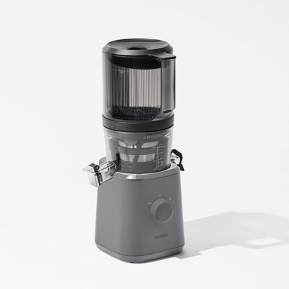 The Nama J2, a modern gray cold press juicer with a transparent top container and front control dial, stands on a white surface, casting a subtle shadow.