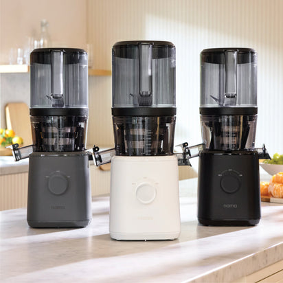 Three Nama J2 vertical cold press juicers in black, white, and dark gray sit on a kitchen counter, with blurred fruits and other kitchen items in the background.