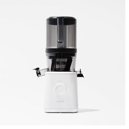 The Nama J2 Plug Type I is a sleek, modern cold press juicer with a white base, large central dial, transparent pulp container, and black accents. It features hands-free juicing and displays the nama logo on the front against a plain white background.