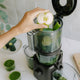 A hand places a halved green apple into the Nama J2 Plug Type I cold press juicer filled with leafy greens, cucumber, and celery. Two glasses of green juice and a plate with cut cucumber are on the white kitchen counter nearby.
