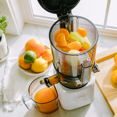 A Nama J2 juicer filled with orange and lime segments sits on a kitchen counter near a window, collecting fresh juice in its container, with assorted citrus fruits arranged on plates and cutting boards nearby.