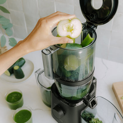A hand places a green apple half into the Nama J2 Plug Type G juicer filled with leafy greens and cucumbers; green juice-filled glasses and a cucumber sit on a white countertop, highlighting the easy-to-clean design.