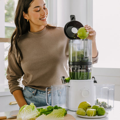 A woman smiles as she puts a green apple into the Nama J2 Plug Type E in a bright kitchen. Fresh apples, lettuce, and leafy greens on the counter highlight its easy-to-clean and hands-free juicing features.